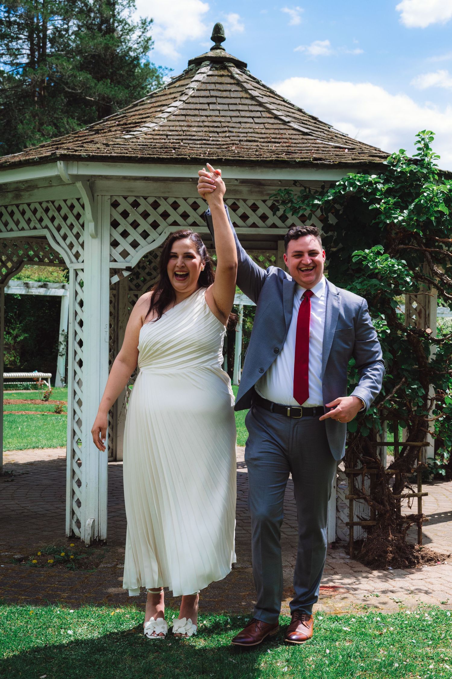 A couple in wedding attire dances and celebrates together in a garden setting with a white gazebo in the background.
