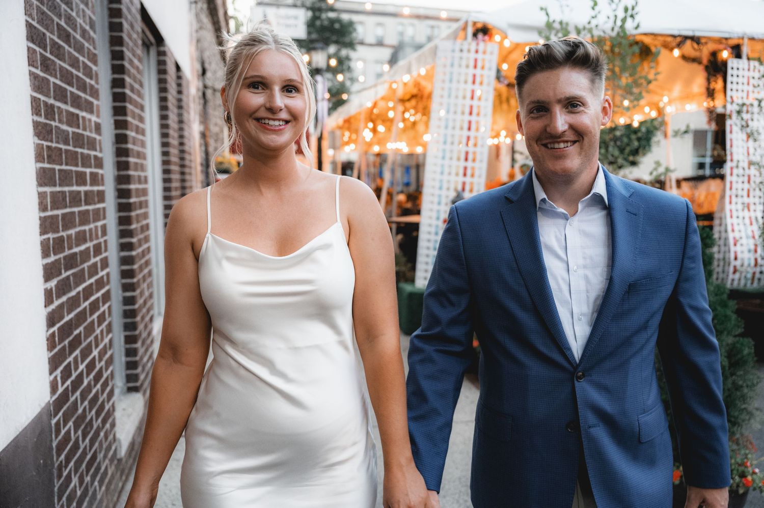 Wedding couple shares a candid moment together while walking along a brick wall in formal attire.