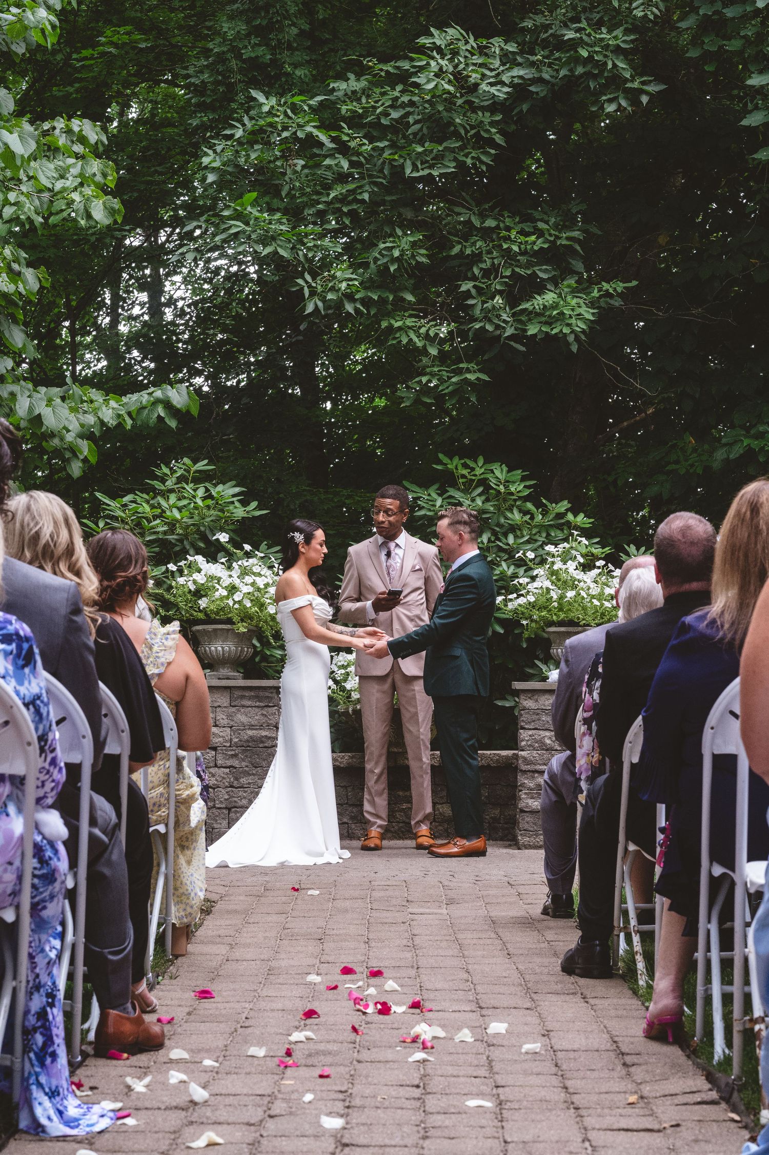 An outdoor wedding ceremony takes place on a brick pathway surrounded by lush greenery and stone walls.