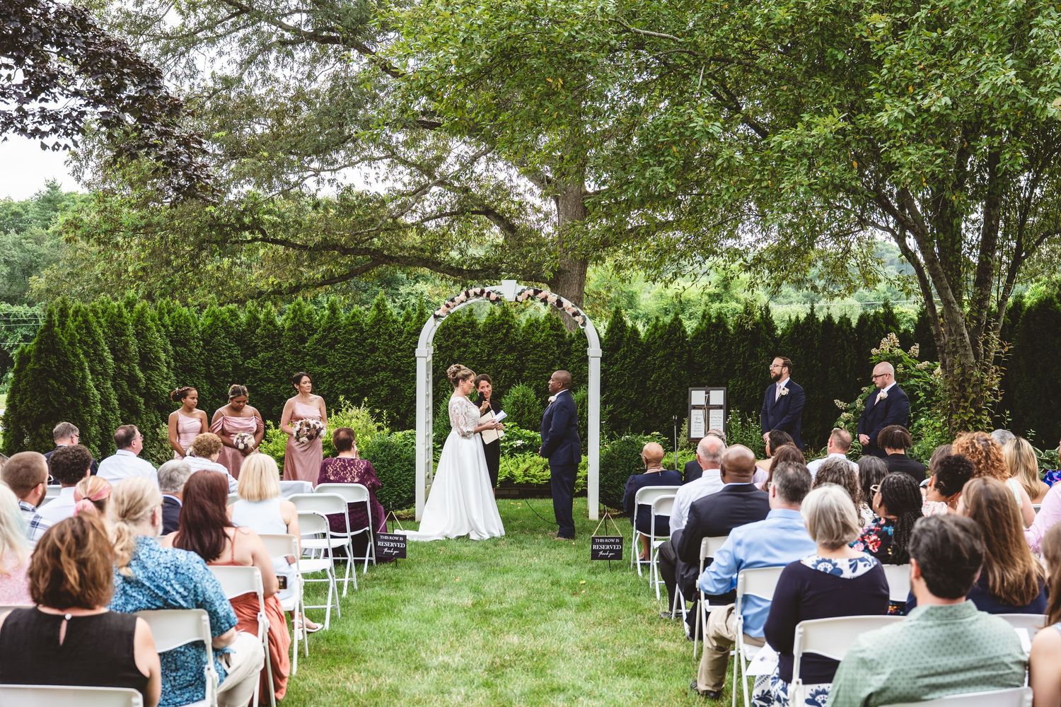 Guests seated in white chairs during an outdoor garden wedding ceremony beneath lush green trees.