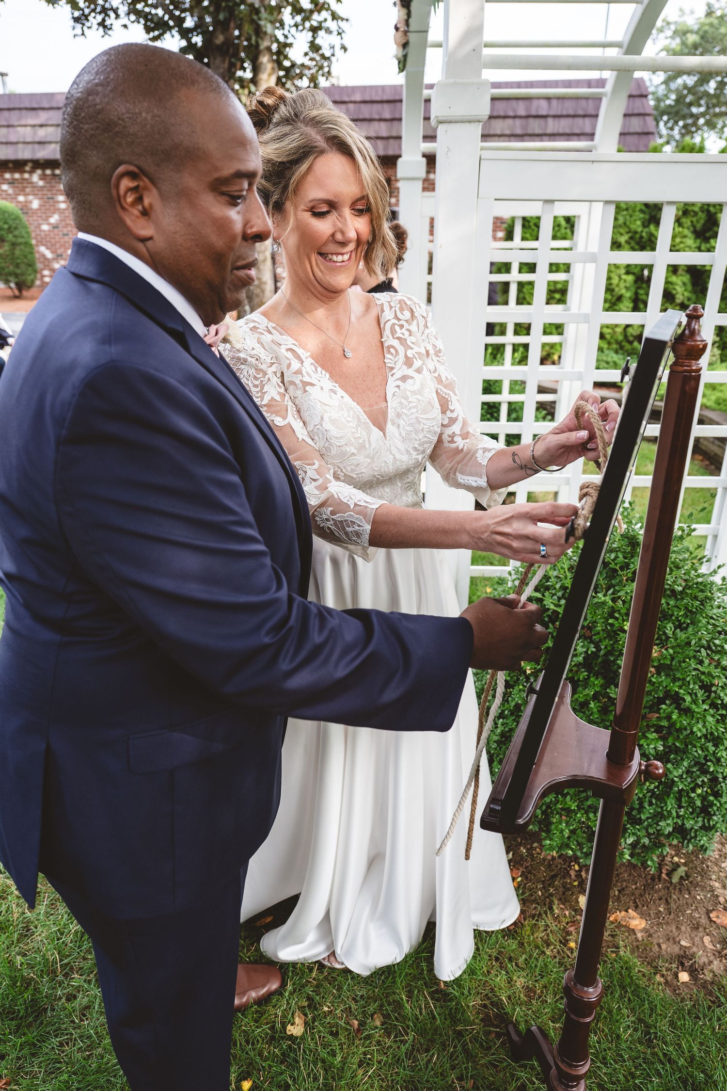 A couple in wedding attire share a playful moment on a wooden bench in a garden setting with white lattice fencing.