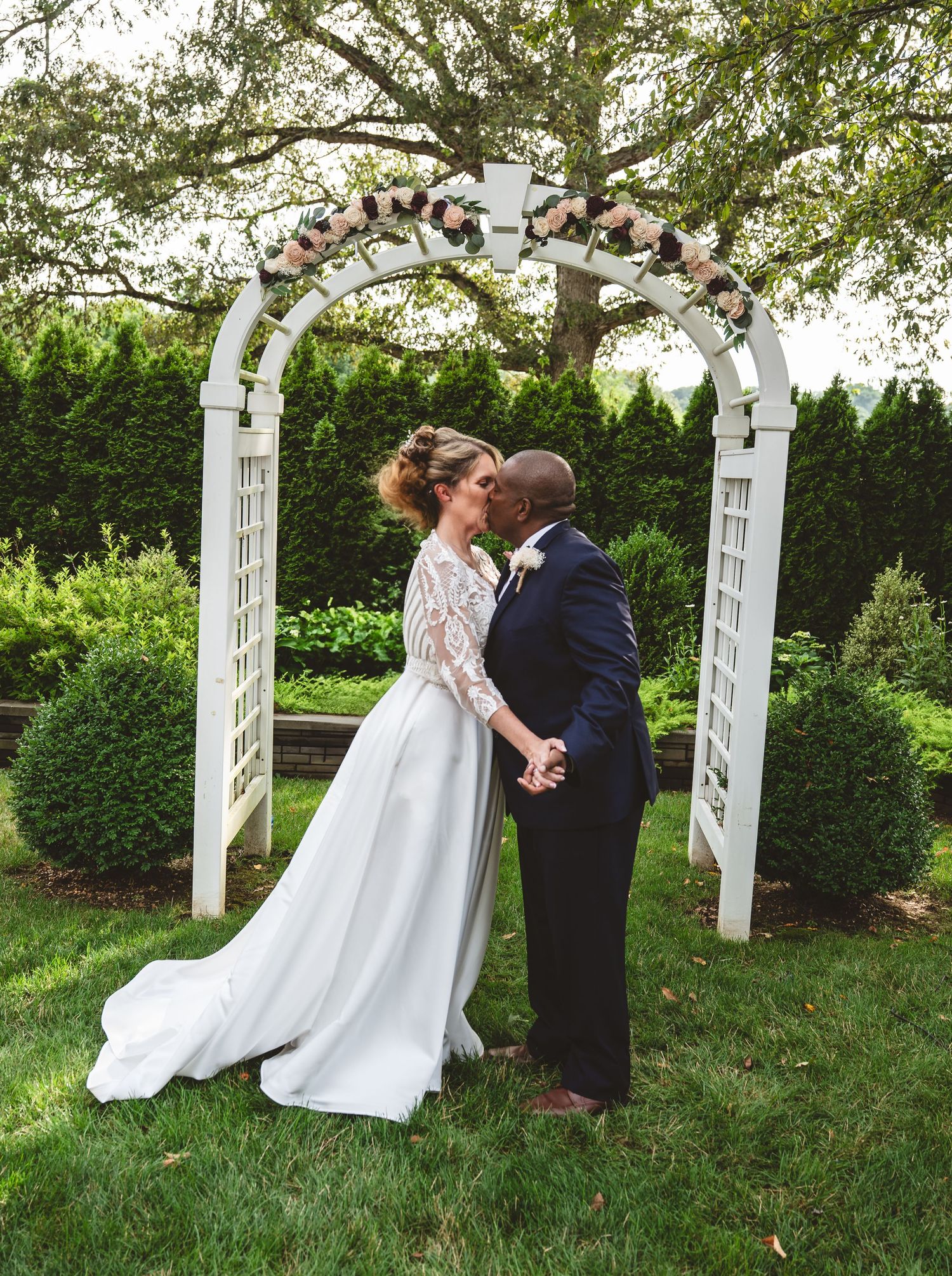 A romantic wedding scene under a white floral arch in a lush garden setting with a flowing white gown and navy suit.