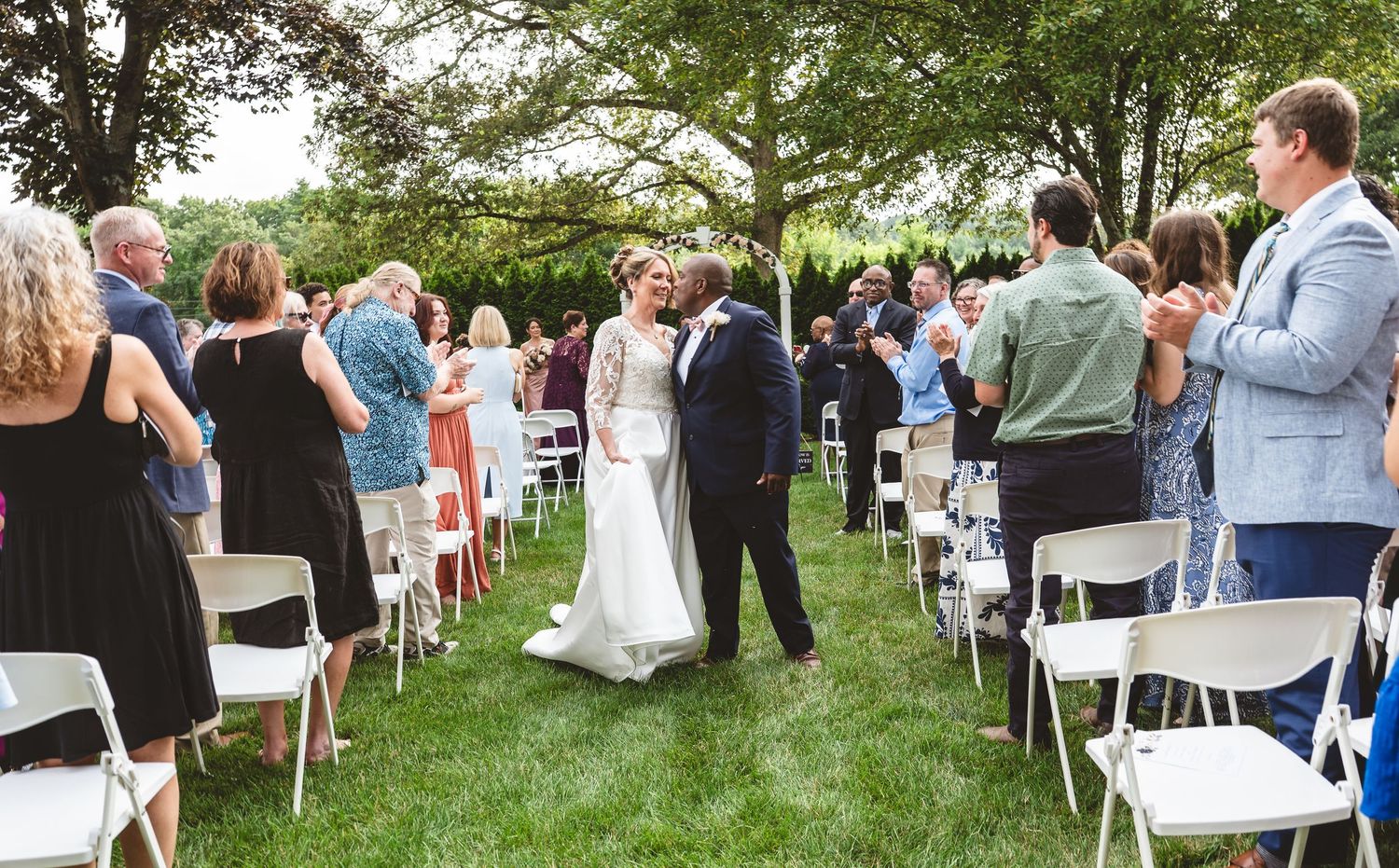 A panoramic view of an outdoor wedding ceremony setup with white chairs arranged on a green lawn under tall trees.
