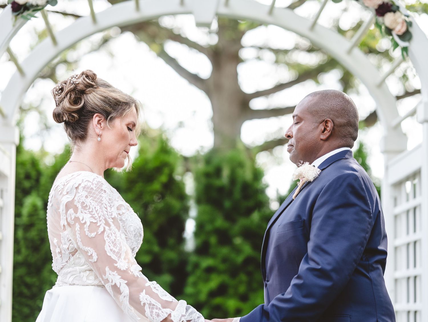 A bride and groom share a tender moment under a white garden arbor during their outdoor wedding ceremony.