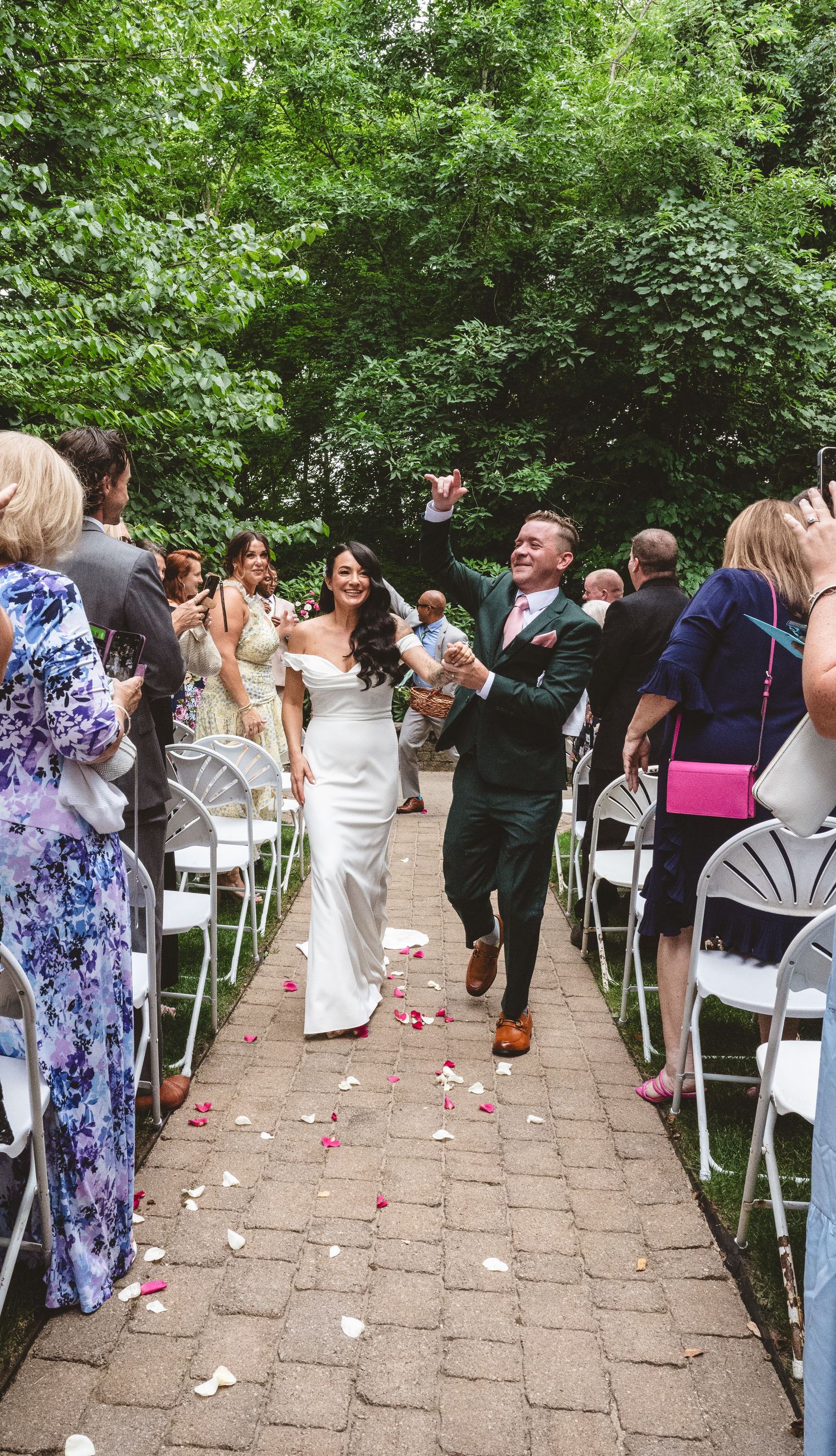 Wedding guests stand and cheer during an outdoor garden ceremony with white chairs and scattered rose petals.