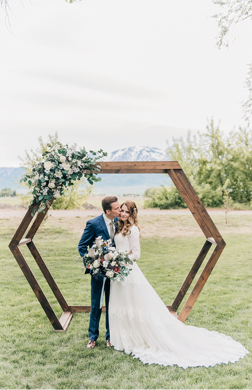 Romantic outdoor wedding ceremony features a wooden hexagonal arch decorated with white flowers against mountain backdrop.