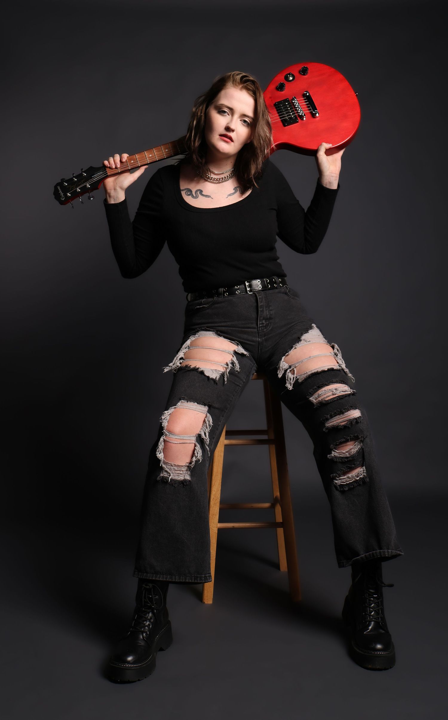 Rock musician in black outfit with ripped jeans poses on stool holding red electric guitar against dark background.