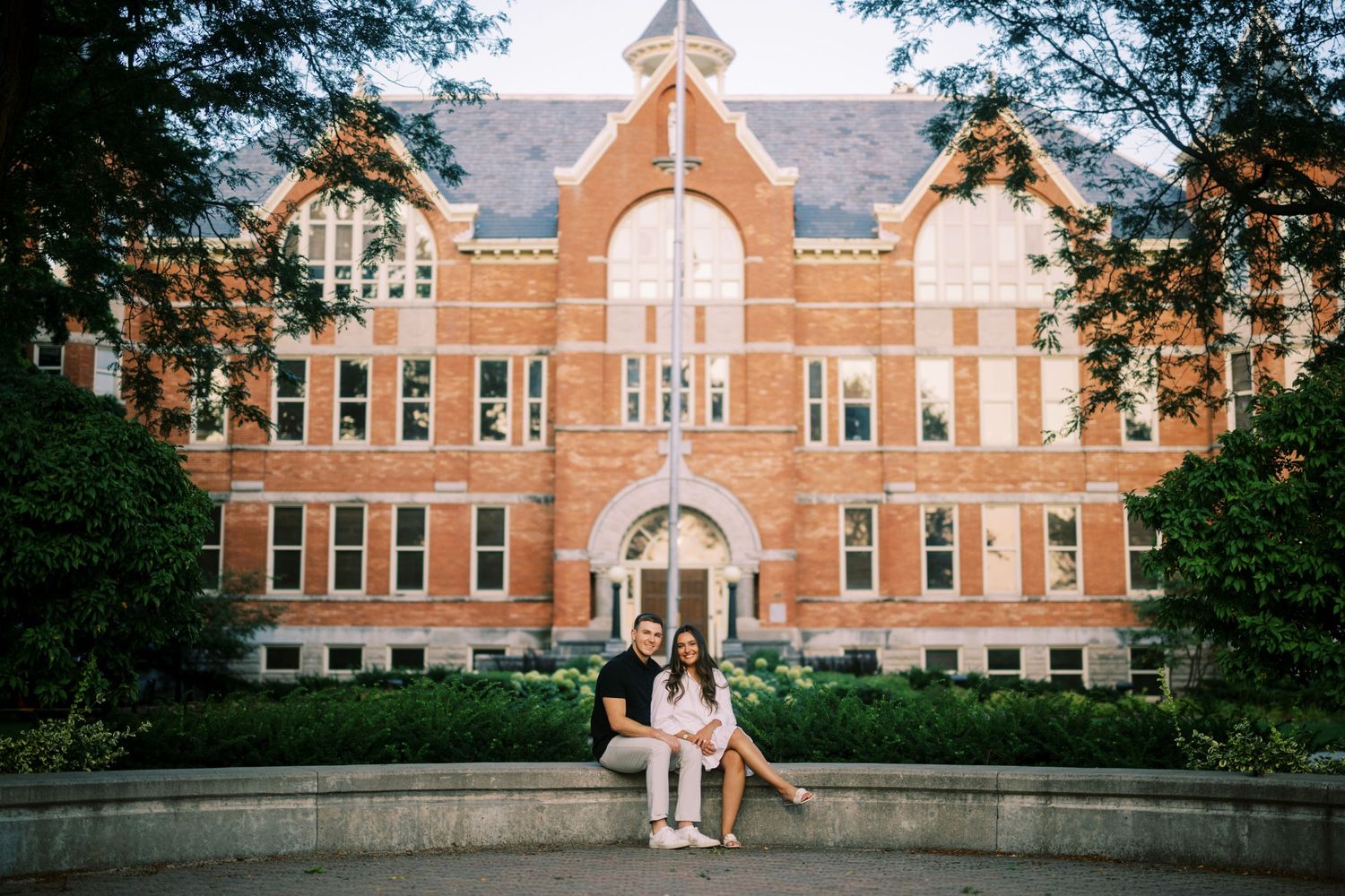 A couple sits on a ledge in front of a grand red brick Victorian Gothic university building with ornate architecture.