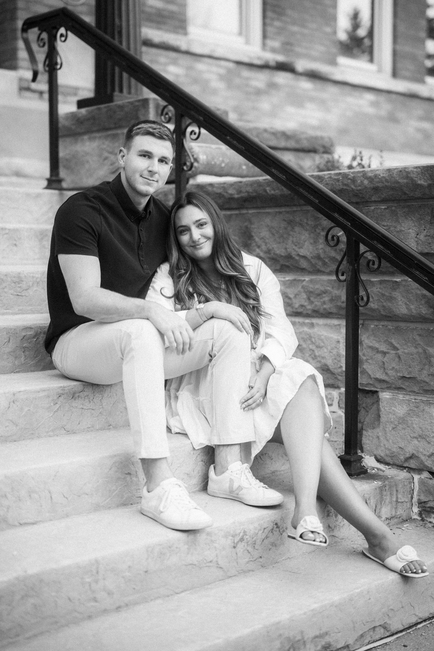 A couple sits close together on stone steps with decorative iron railings in a black and white portrait.