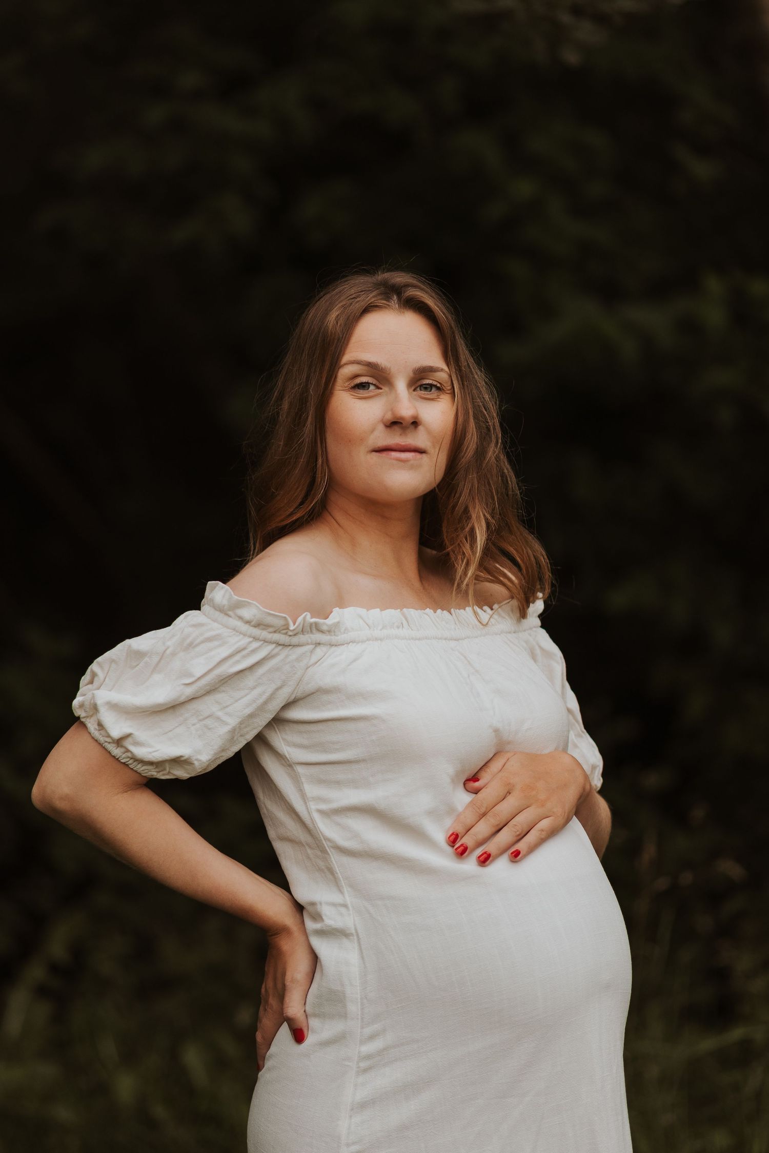 A pregnant woman in a white off-shoulder dress stands in a natural outdoor setting with trees and grass.