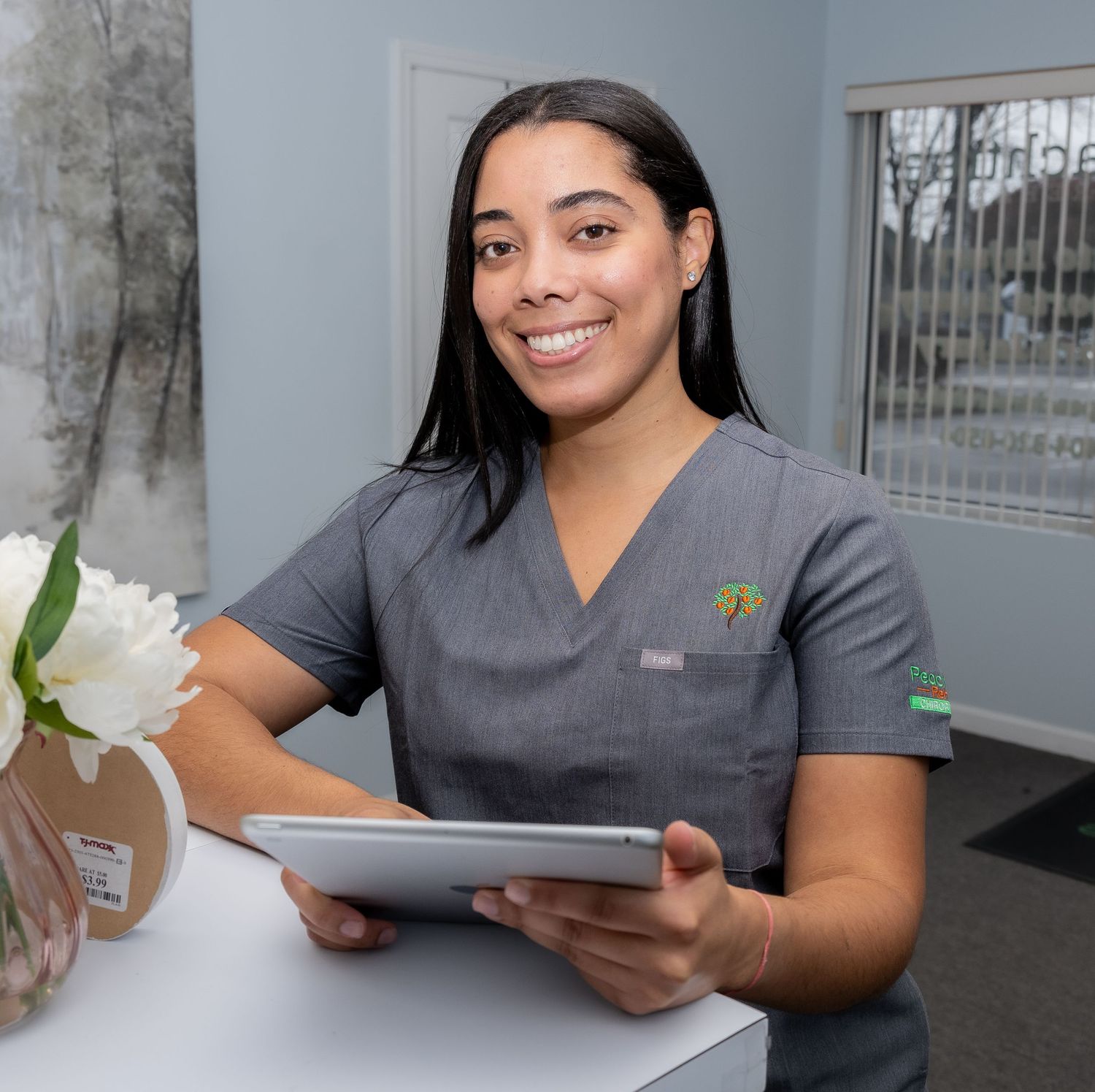 Healthcare professional in gray scrubs smiles while holding a digital tablet at medical office desk with white flower.