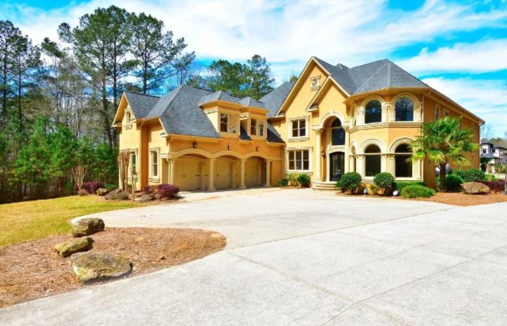 Luxurious yellow stucco mansion with three-car garage and circular driveway surrounded by pine trees in an upscale neighborhood.