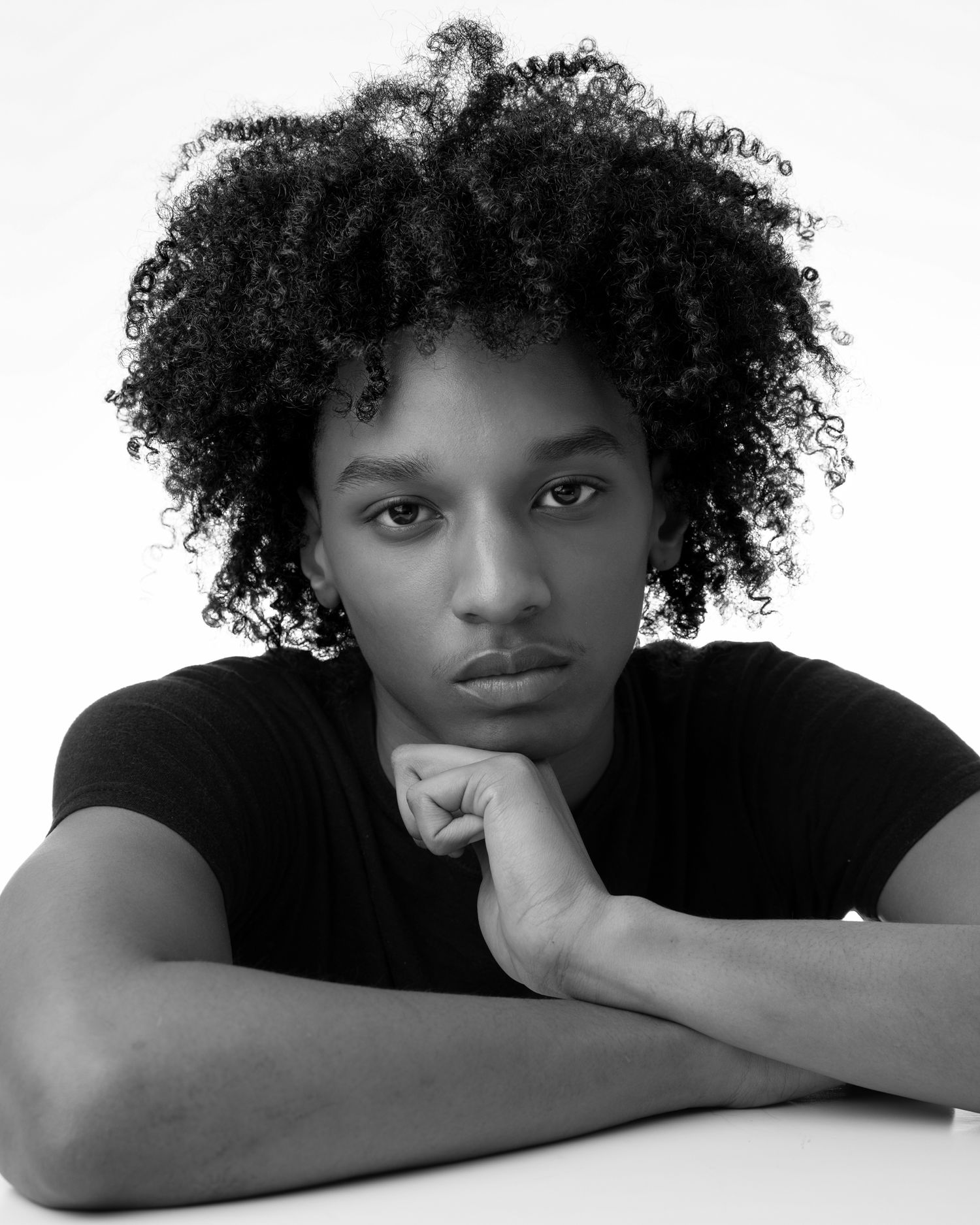 Black and white portrait showing someone in a black shirt with natural curly hair resting their chin on their hand.