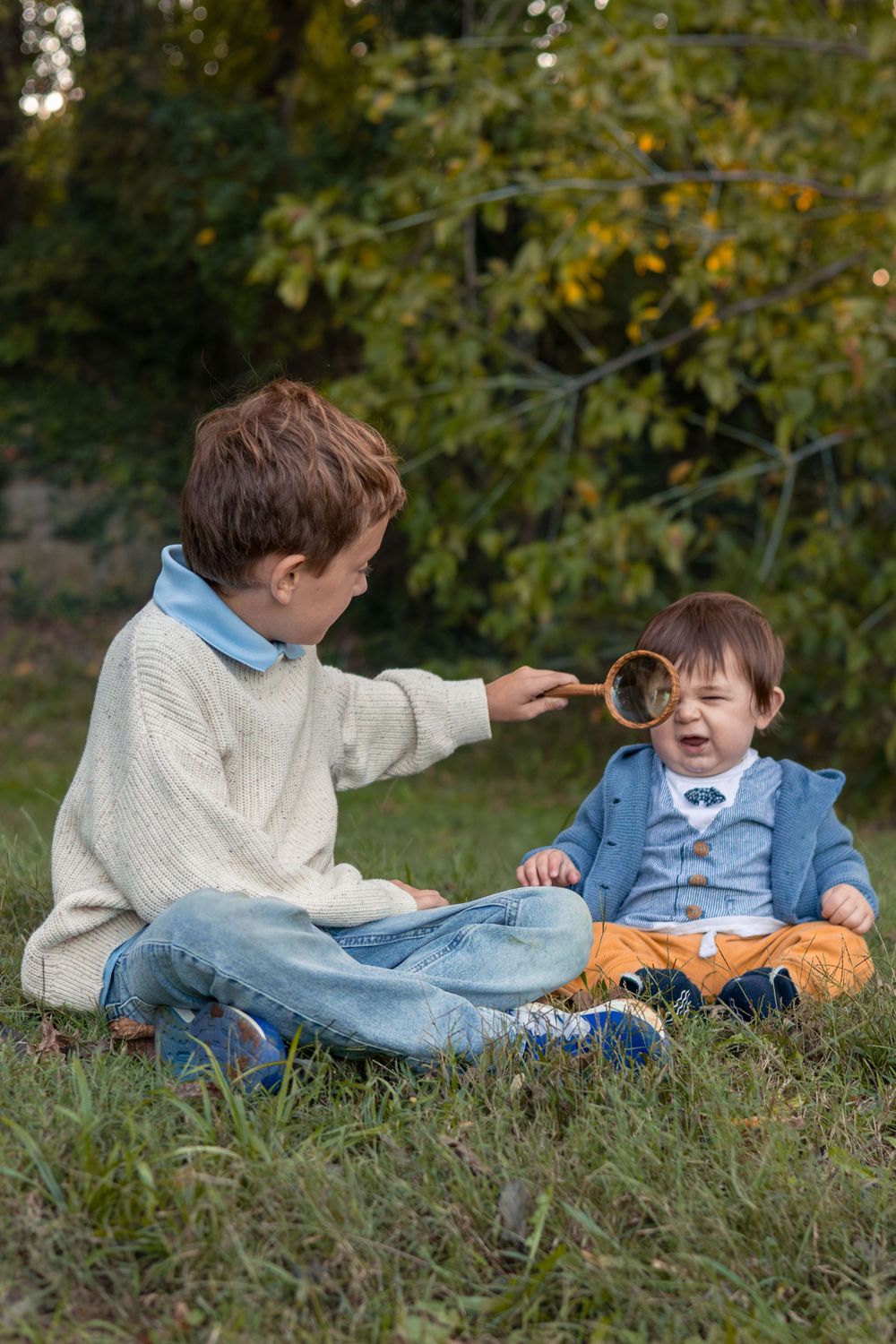 A young boy holds a magnifying glass to his toddler brothers eye.