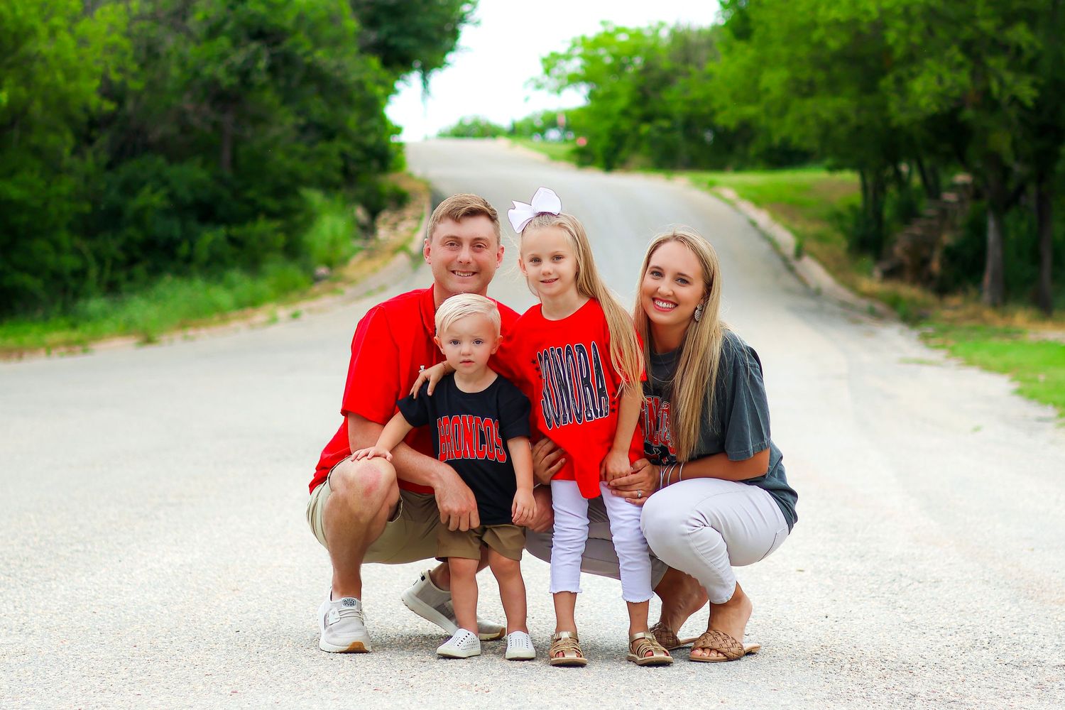 A group crouches together for a casual photo on a rural road surrounded by green trees.