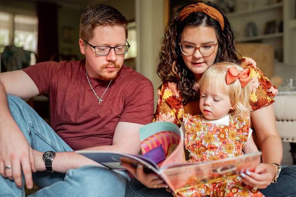 Two adults and a toddler sit together on a couch reading a book in a cozy living room setting.