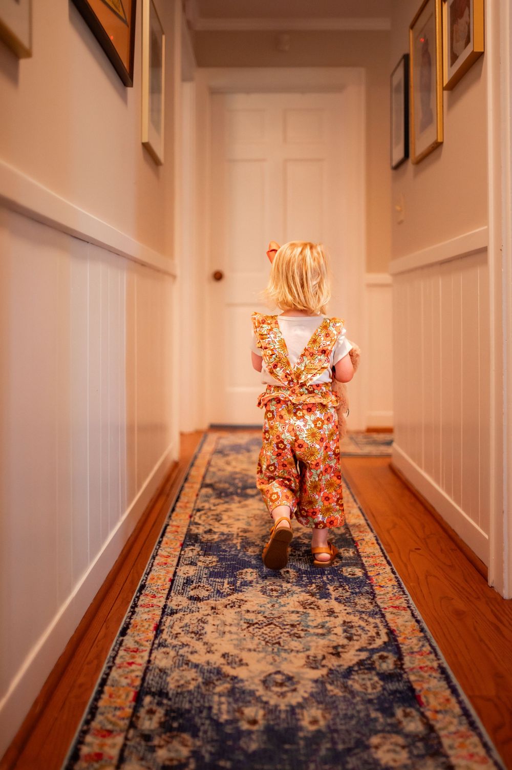Small child wearing floral pajamas runs down a hallway with hardwood floors and a blue runner rug.