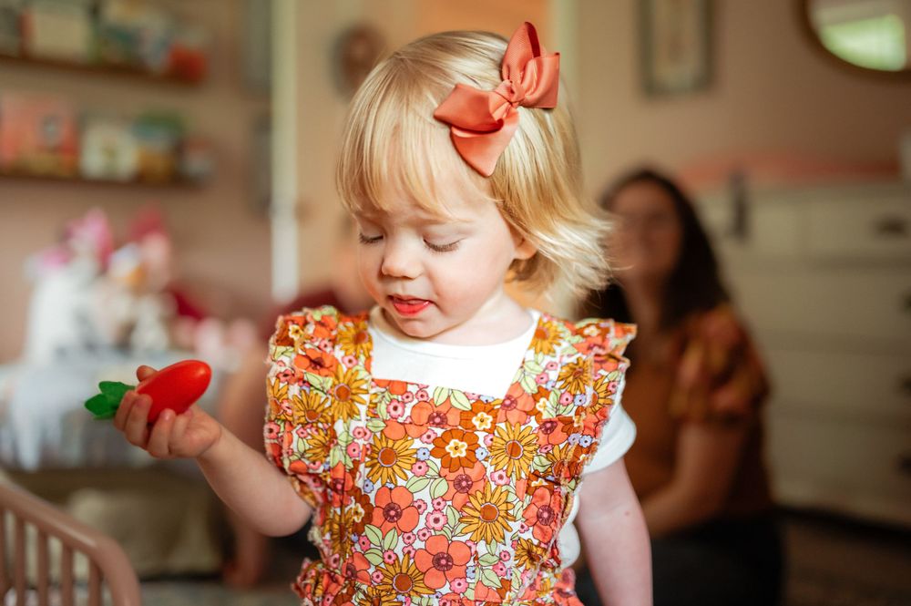 Small child wearing floral dress with red bow plays with red toy in kitchen setting.