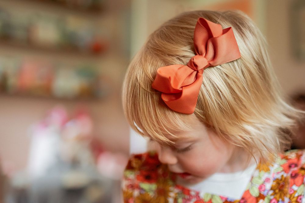 Small child with blonde hair wearing a peach-colored bow and floral dress looks down in soft focus photograph.