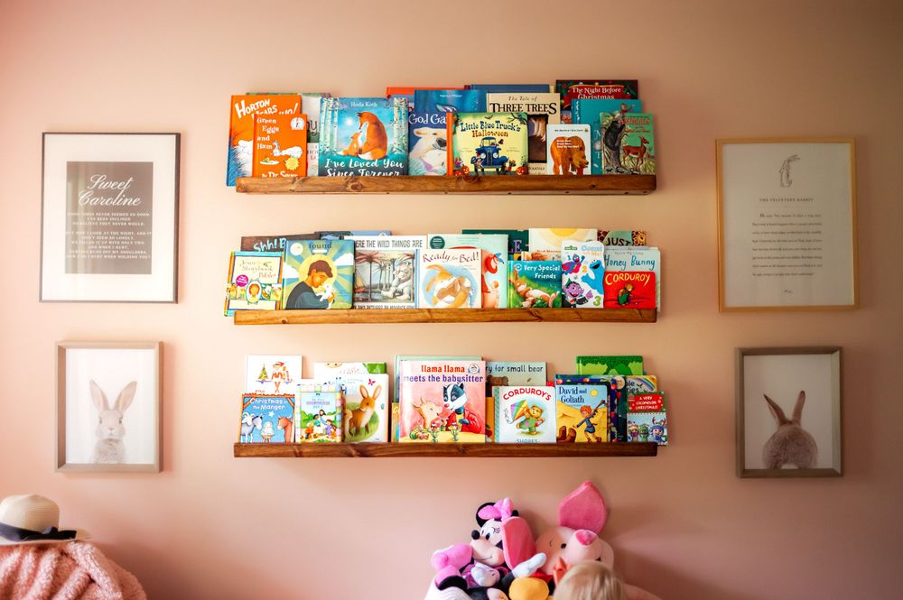 Children's book display on floating wooden shelves with bunny artwork and stuffed animals in a cozy pink nursery corner.