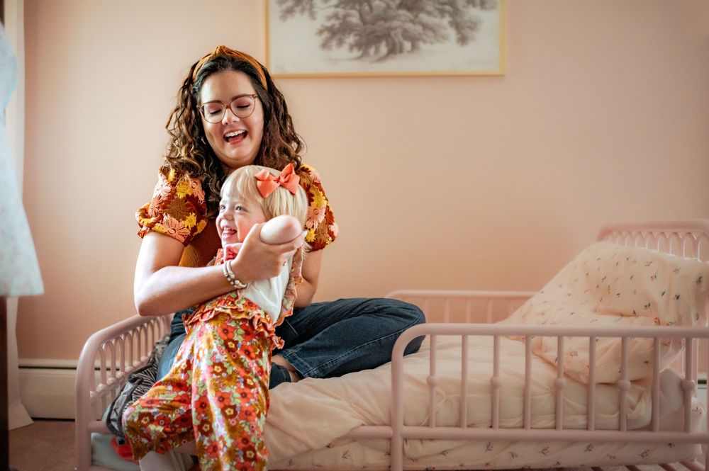 Someone in a floral dress sits on a pink couch in a nursery holding a baby.