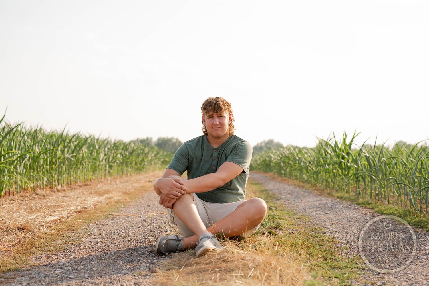 Person in green shirt sits cross-legged on a dirt path between corn fields.