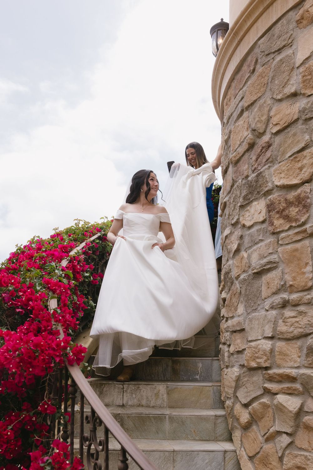 A bride in an elegant off-shoulder white gown descends stone steps beside a stone wall adorned with vibrant red flowers.