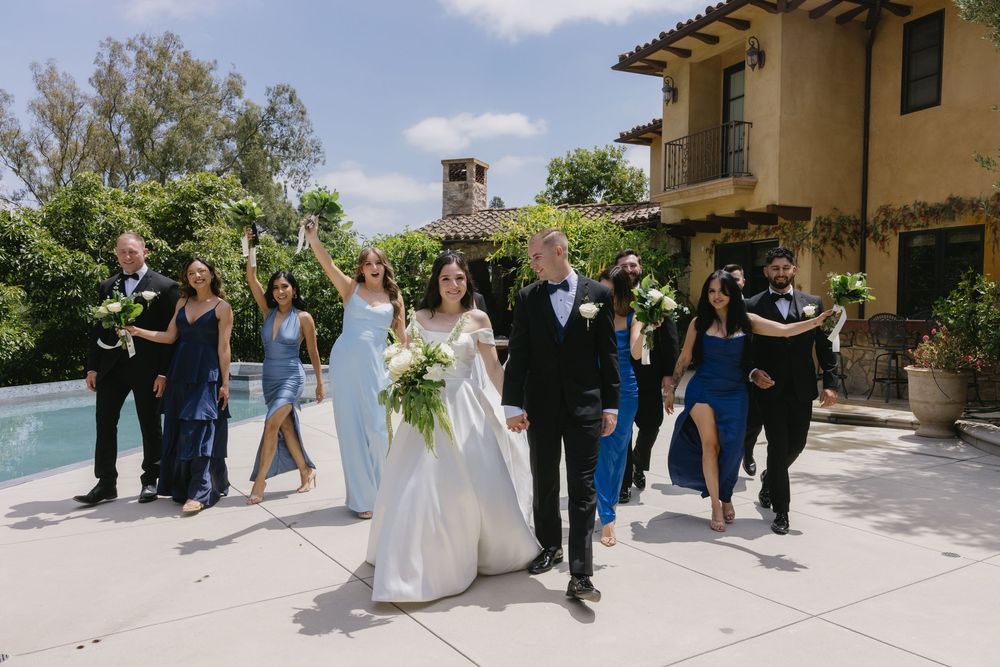 A wedding party walks together and smiles while exiting a Mediterranean-style venue on a sunny afternoon.