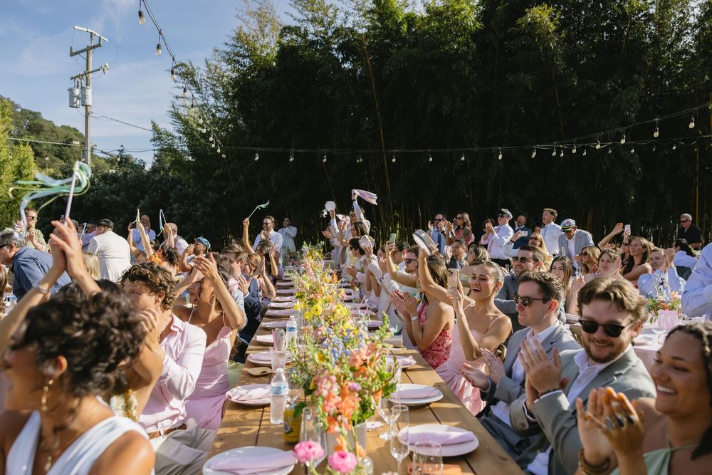 Outdoor wedding reception with guests raising hands at long decorated table with colorful flowers under string lights.
