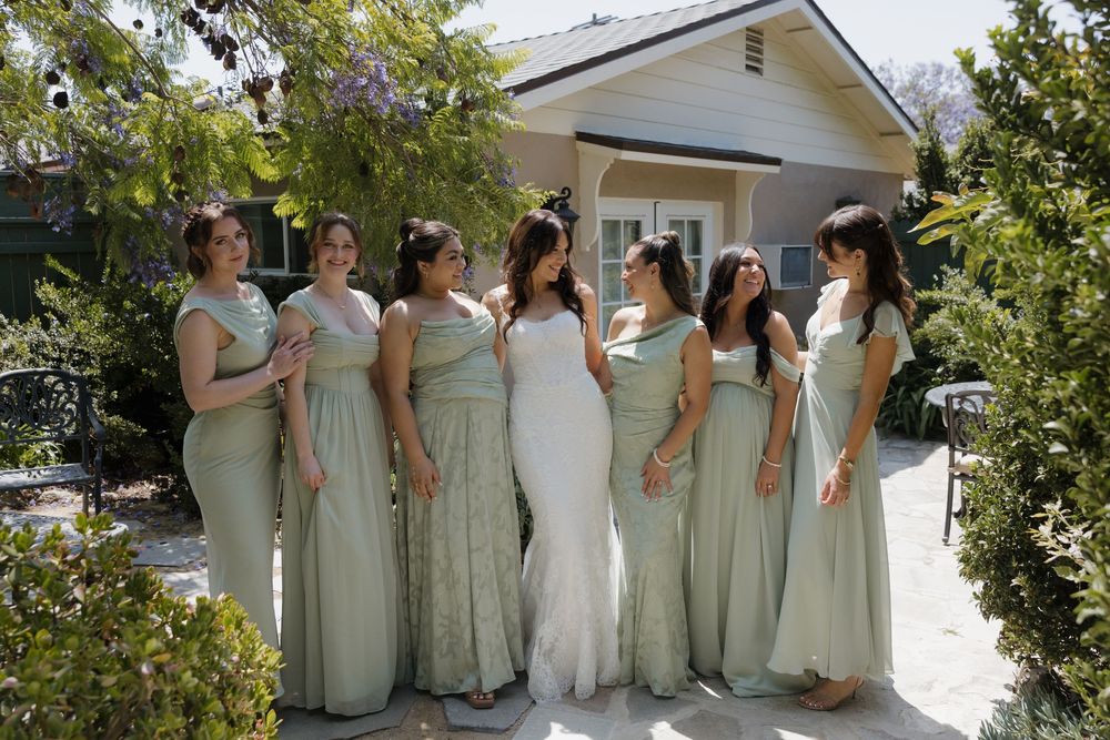 Bride in white gown stands with six bridesmaids in sage green dresses outside a charming house.