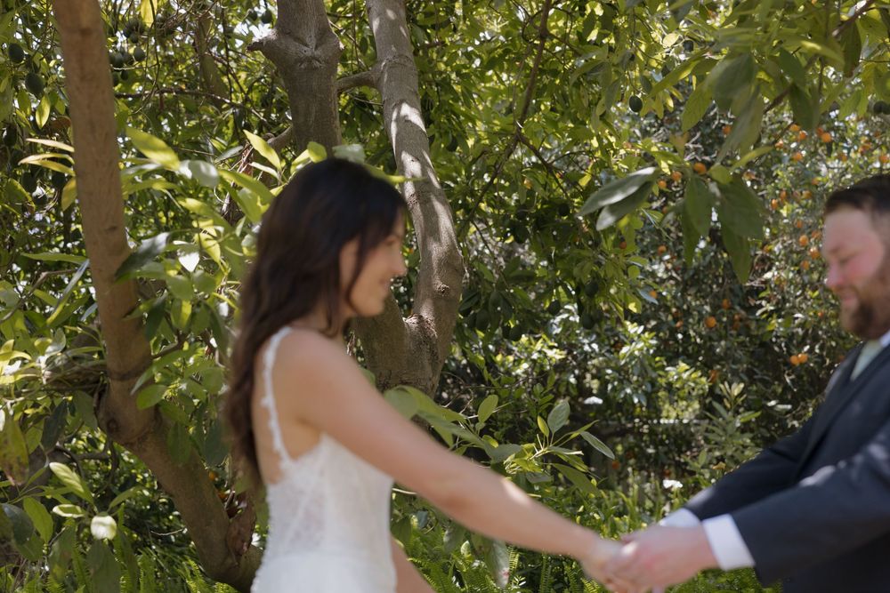 A bride and groom hold hands outdoors surrounded by lush green trees on their wedding day.