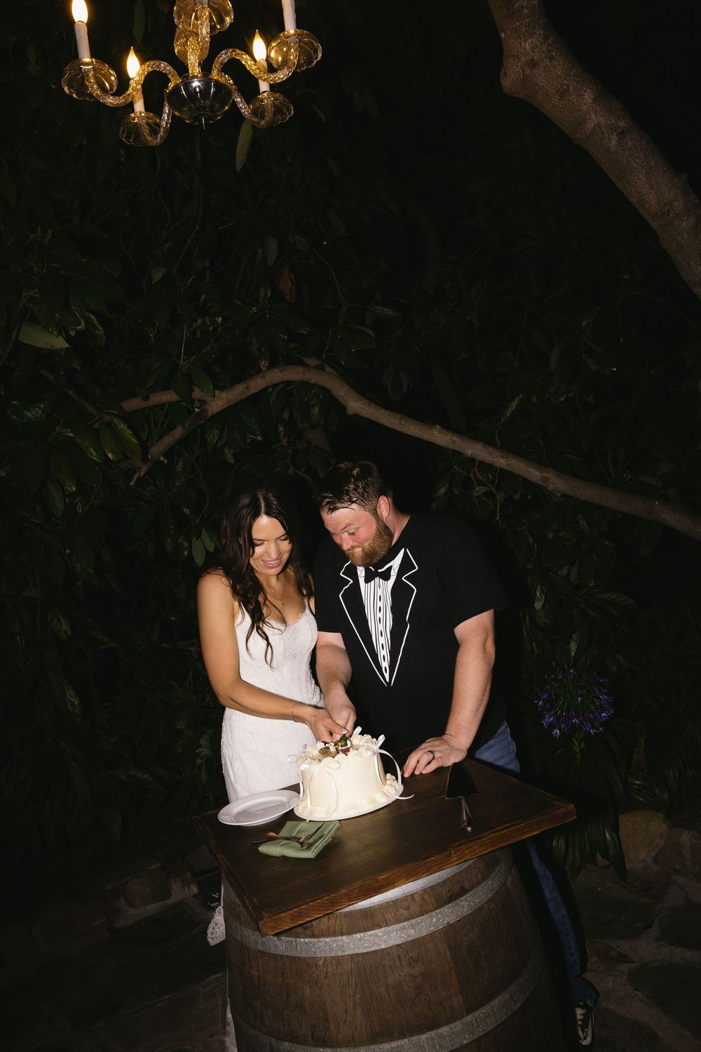 Bride and groom cutting wedding cake on barrel under chandelier at night outdoor ceremony.