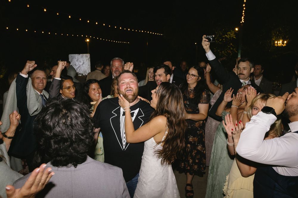 Bride and groom dancing joyfully with wedding guests at an outdoor evening reception under string lights.