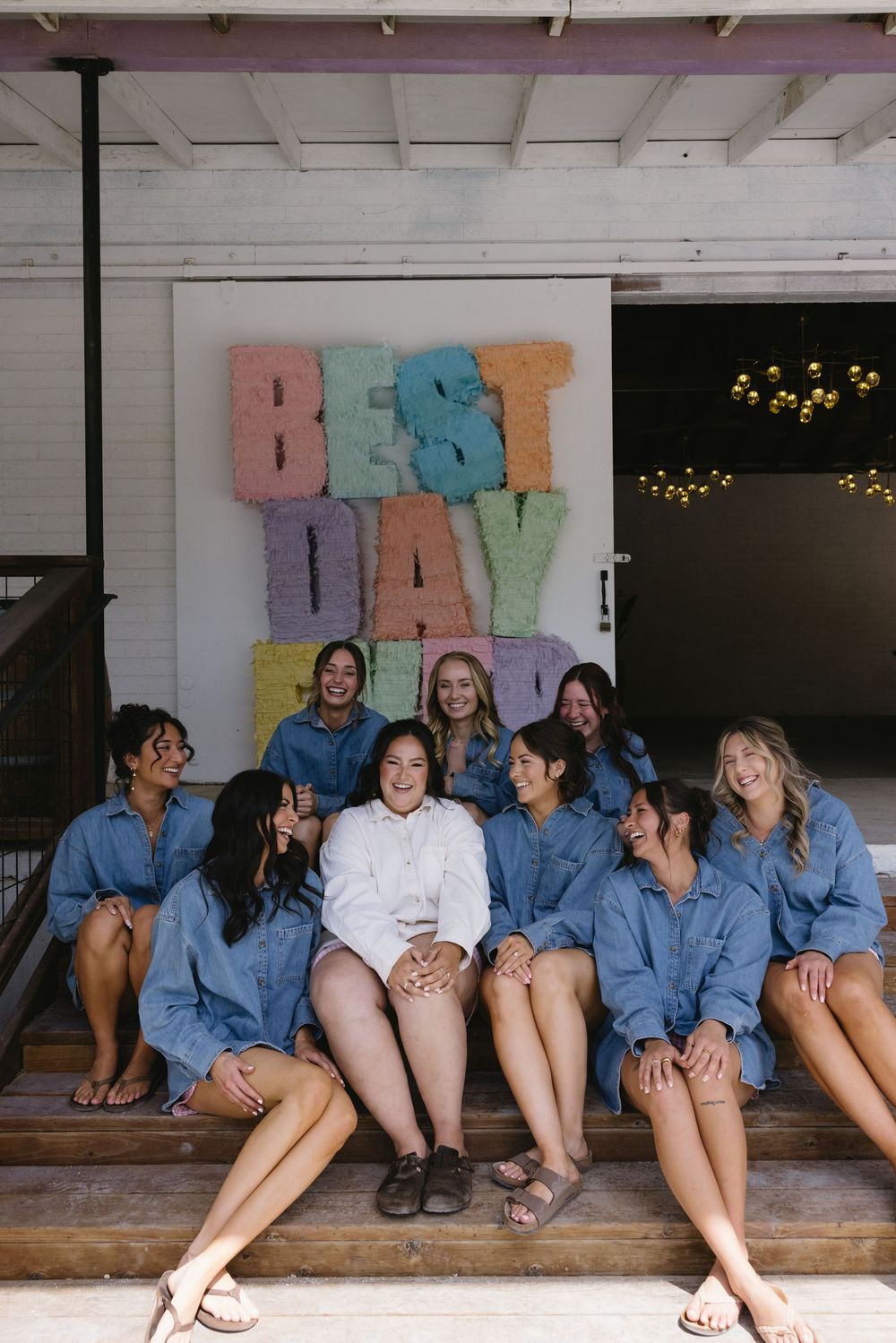 Bride in white with bridesmaids in denim sitting on steps in front of colorful Best Day Ever letter backdrop.