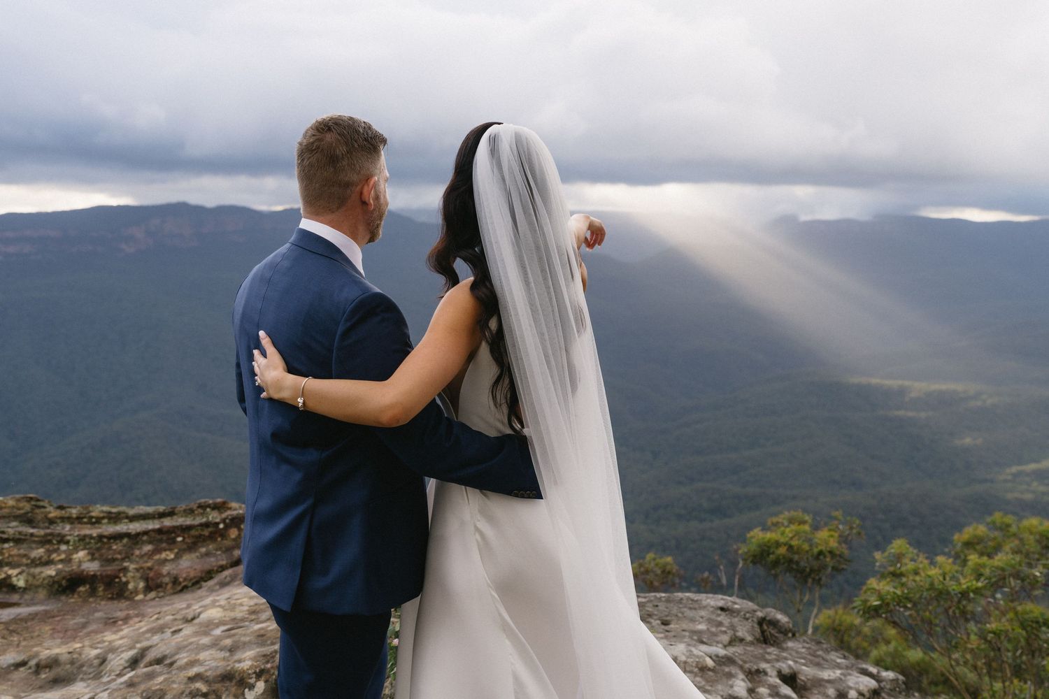 Couple stands together on mountain summit overlooking misty valley with dramatic sunbeams breaking through clouds.