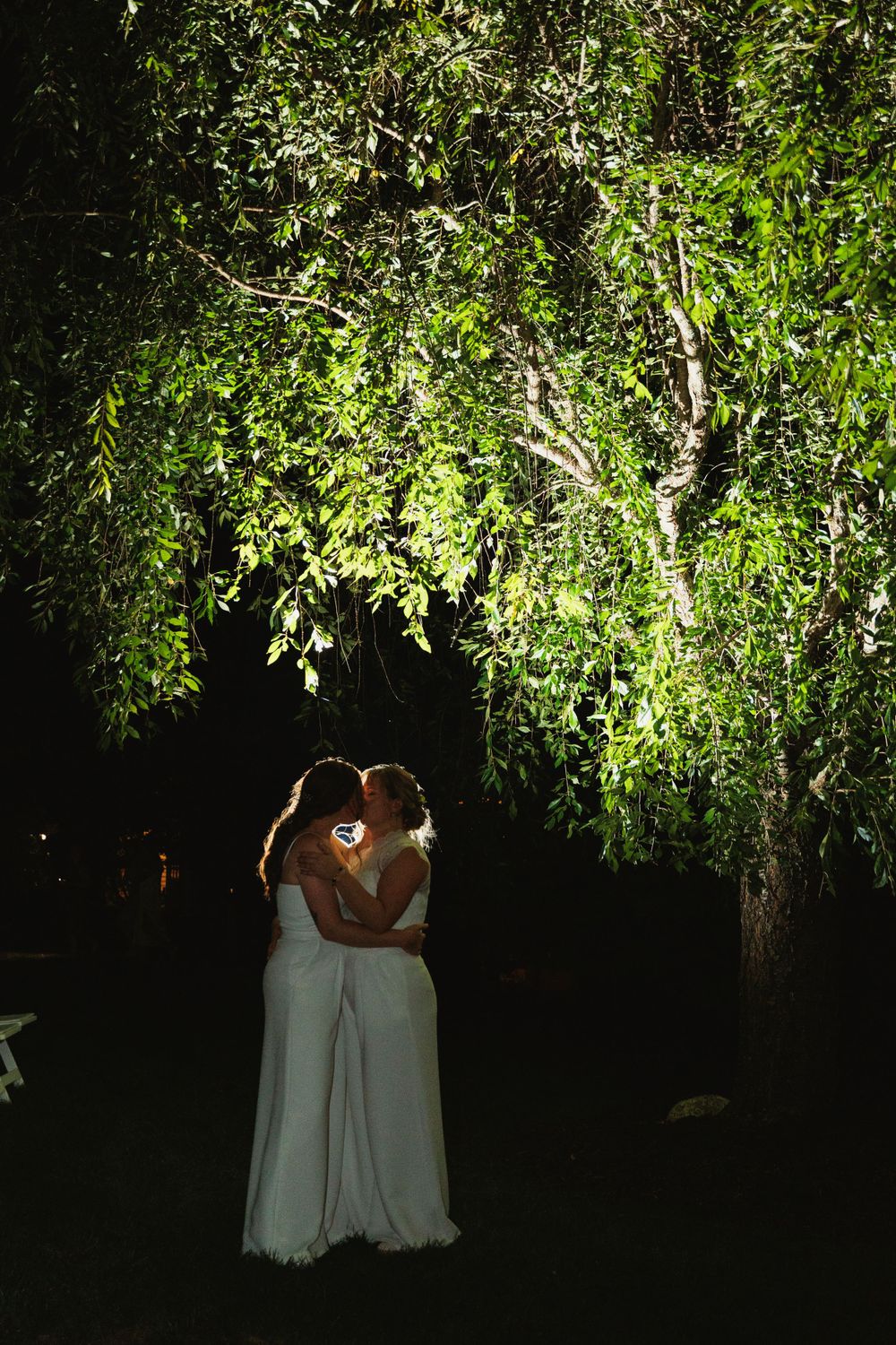 A romantic wedding couple embraces under illuminated tree branches at night.