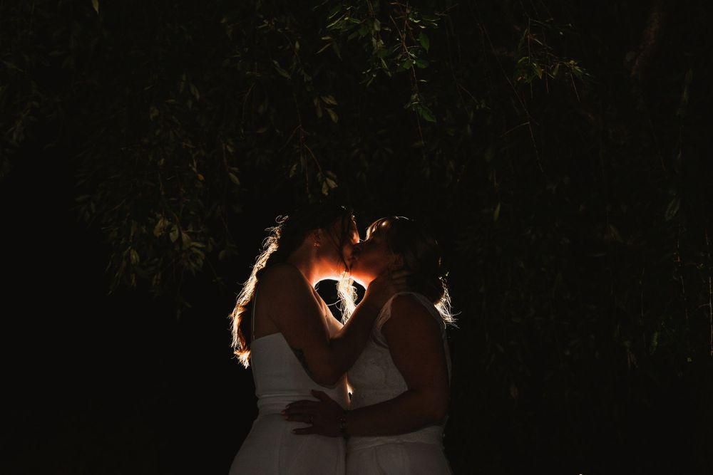 A backlit silhouette of two people in white dresses creating a heart shape with their hands.