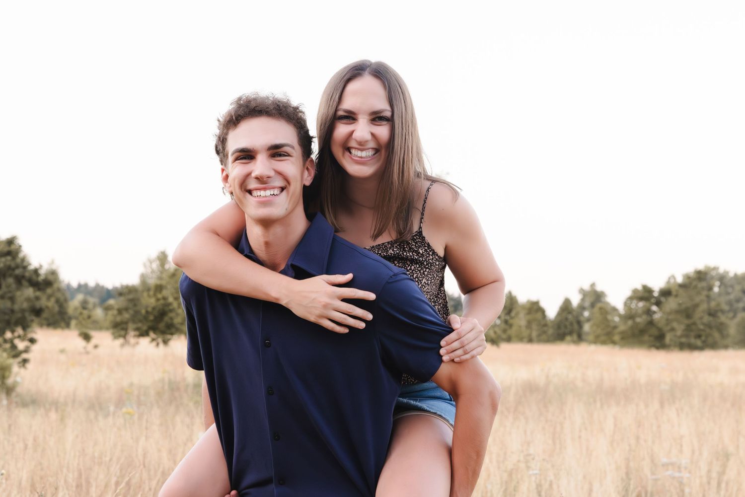 A happy couple shares a piggyback ride moment together in a sun-lit field during golden hour.