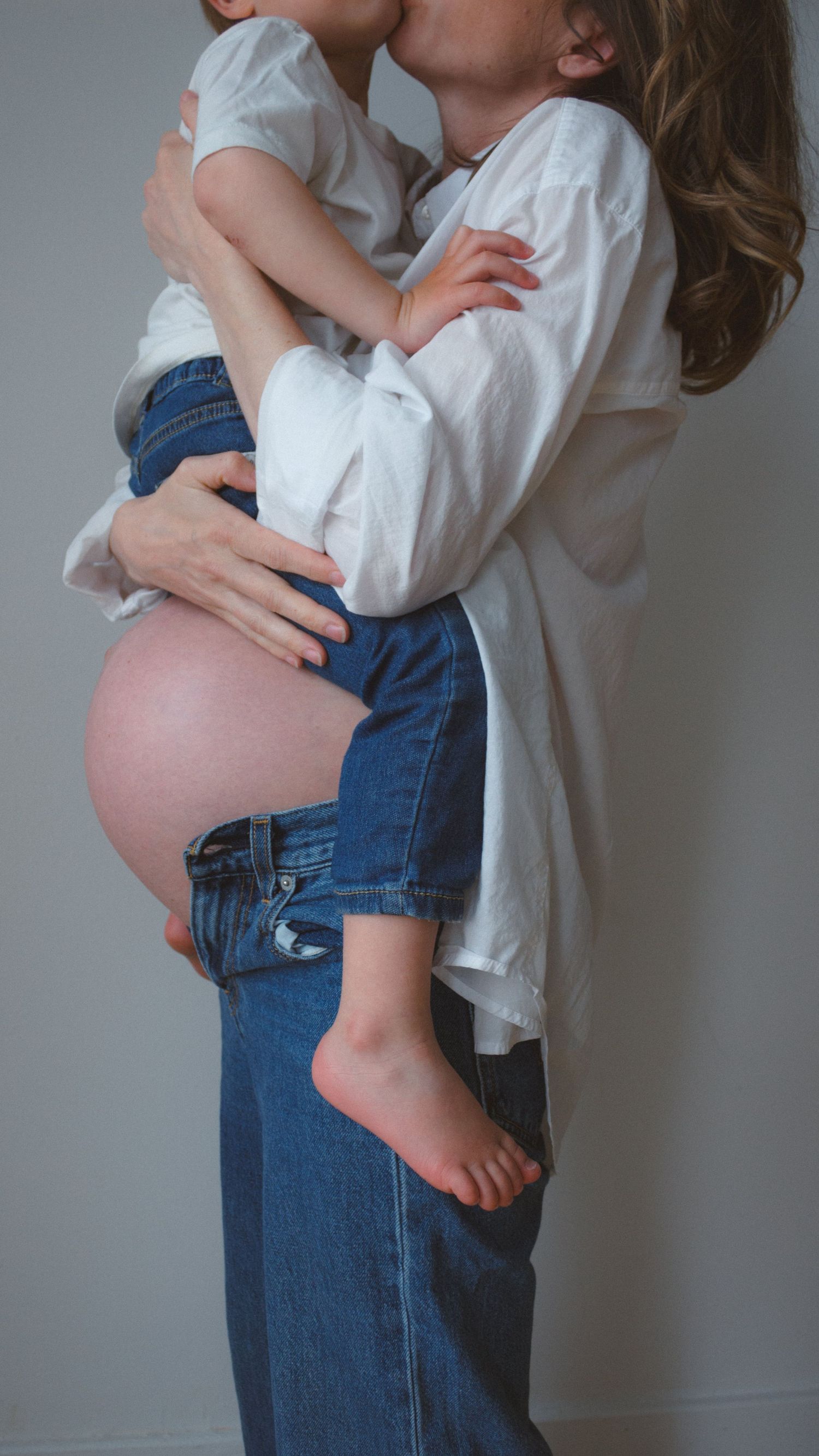 Mom holding toddler over her pregnant belly while giving him a gentle kiss on the cheek during a family maternity session.
