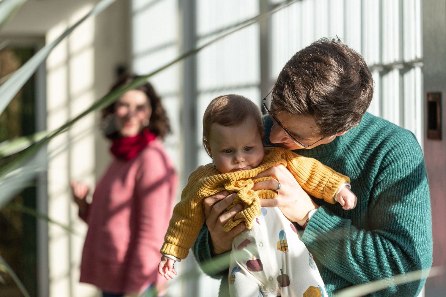 Sunlit indoor family scene with baby in yellow sweater being held near bright windows.