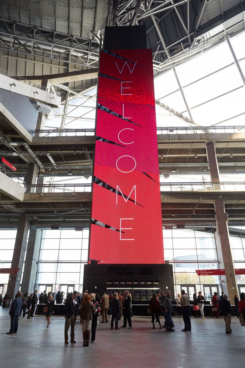 Large red welcome sign pillar in modern stadium concourse with visitors below.