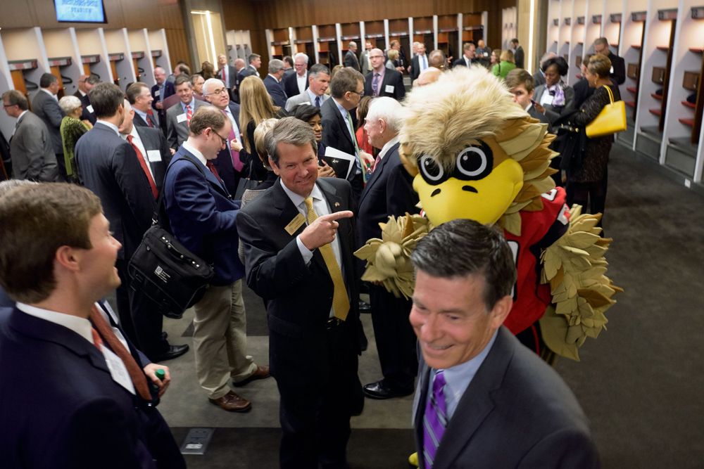 Crowd of people mingling in stadium locker room area with team mascot.