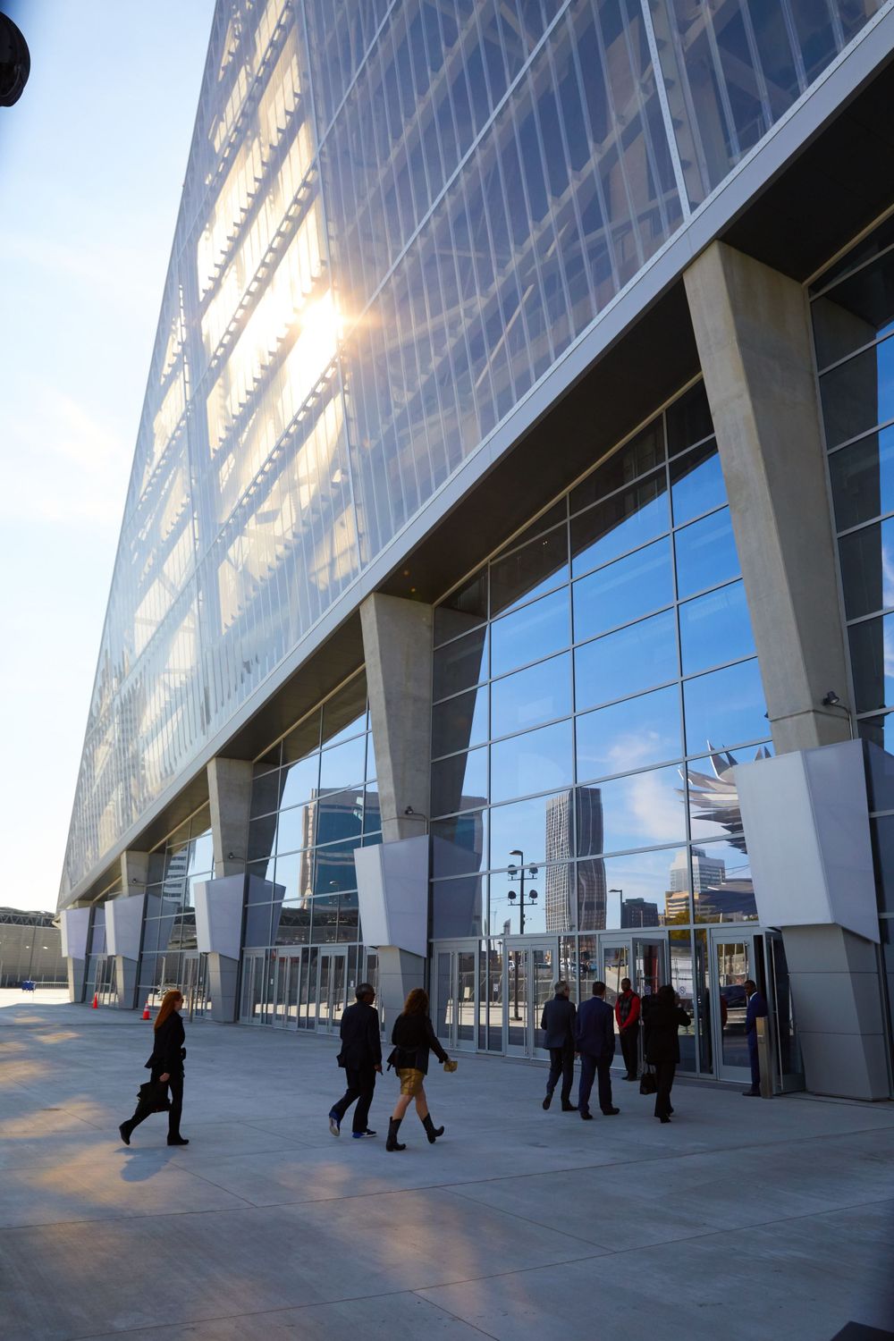 Modern glass exterior of Mercedes-Benz Stadium with people walking underneath at sunset.