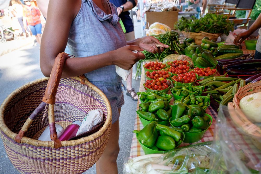Shopper with woven basket browsing fresh produce and vegetables at an outdoor farmers market.