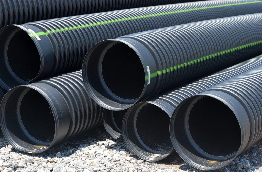 Stack of large black corrugated drainage pipes with green stripes lying on gravel.