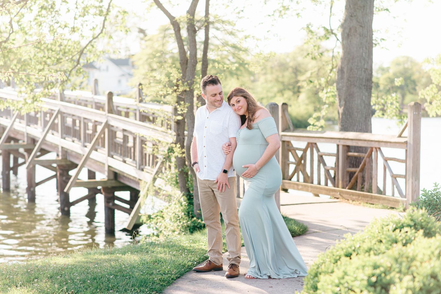 Parents to be stand in front of a sunny bridge poses for maternity photos.