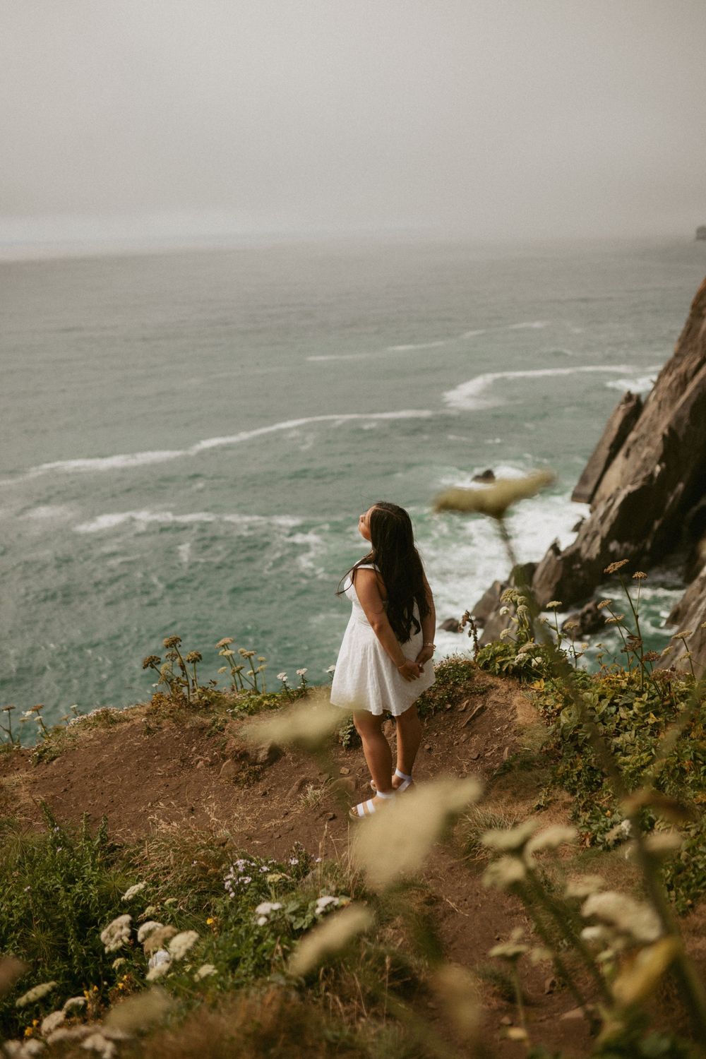People stand on misty coastal cliffs overlooking dramatic waves crashing against rocky shores in overcast weather.