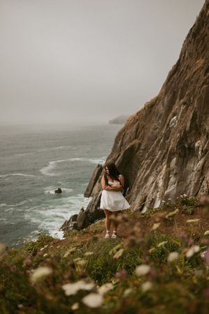 Person in a white dress stands on rugged coastal cliff overlooking misty ocean waves and dramatic rocky shoreline.
