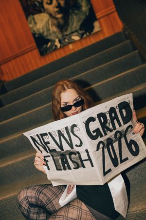 Young woman in sunglasses holding a newspaper reading News Flash Grad 2026 while sitting on stairs.