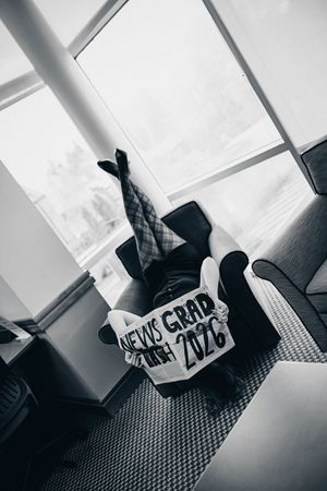 Black and white photo of person sitting in chair holding a sign reading NEWS GRAD 2026.